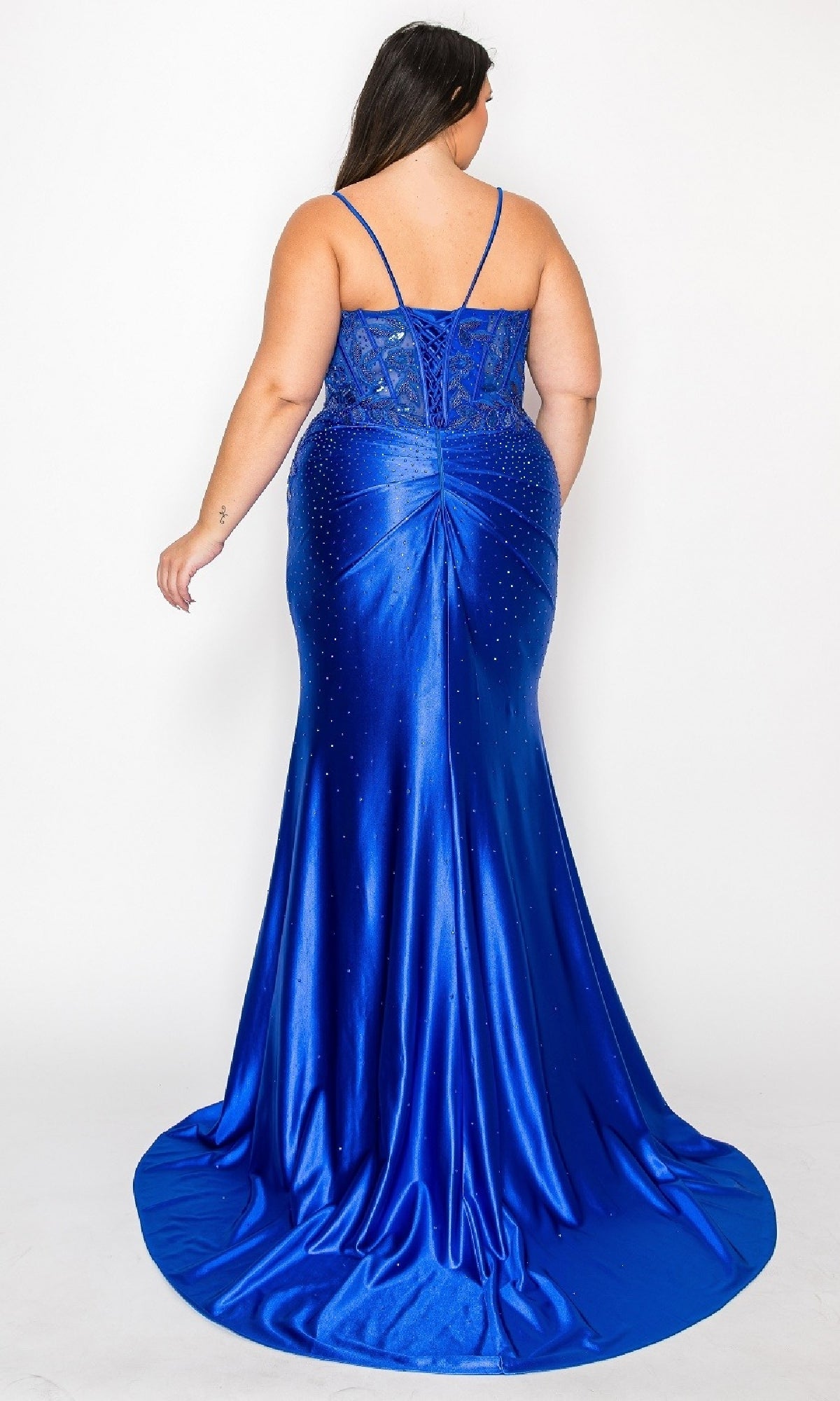 A woman faces away from the camera in the Plus-Size Long Satin Prom Dress (Poly USA 9734), a royal blue gown with thin straps, a lace bodice, rhinestone details, and a flowing floor-length train against a white background.